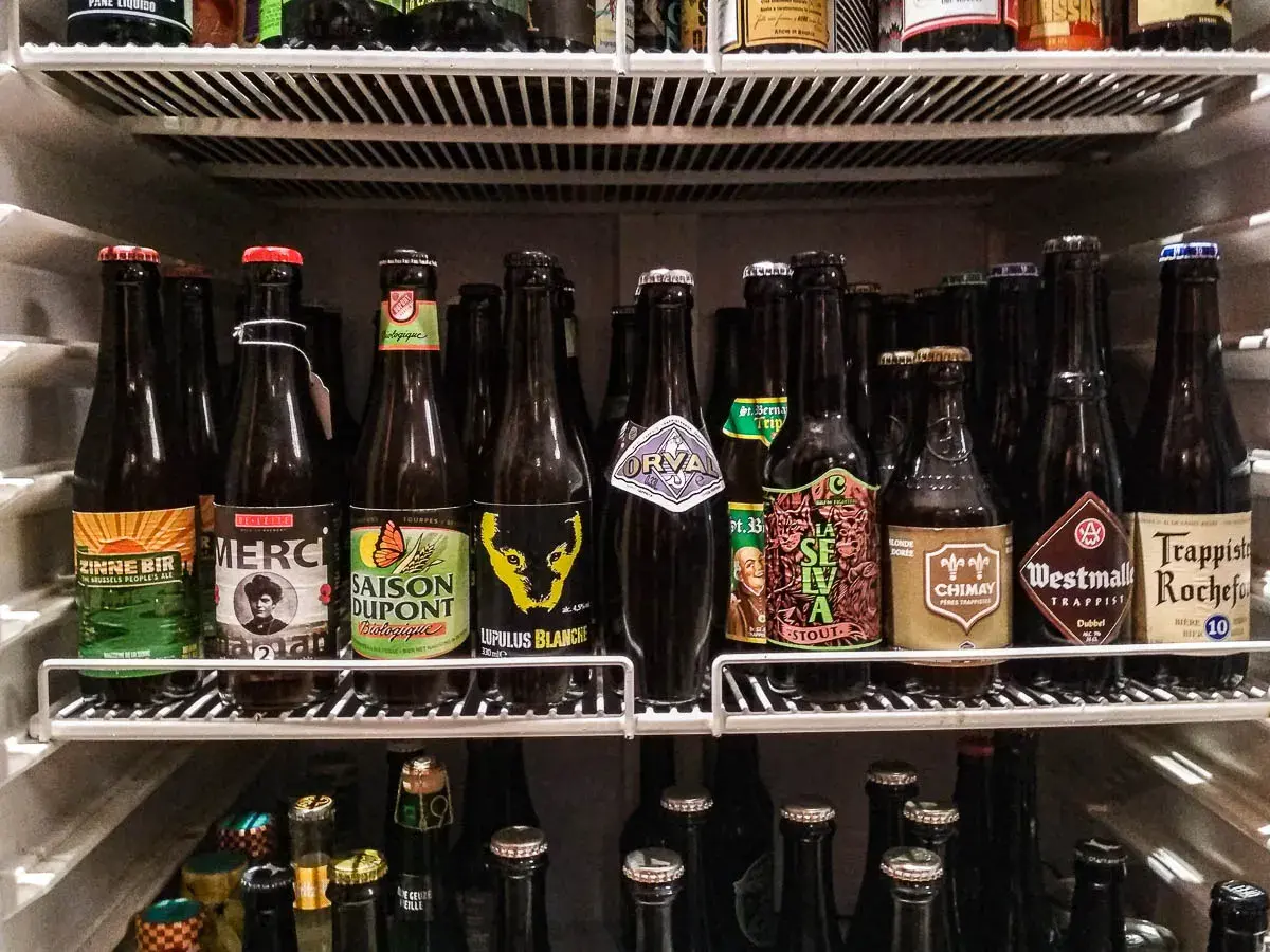 Scaffale pieno di bottiglie di birra, alcune con etichette colorate come "Zinnebir" e "Saison Dupont". Una birra aperta in frigo, pronta per essere gustata.