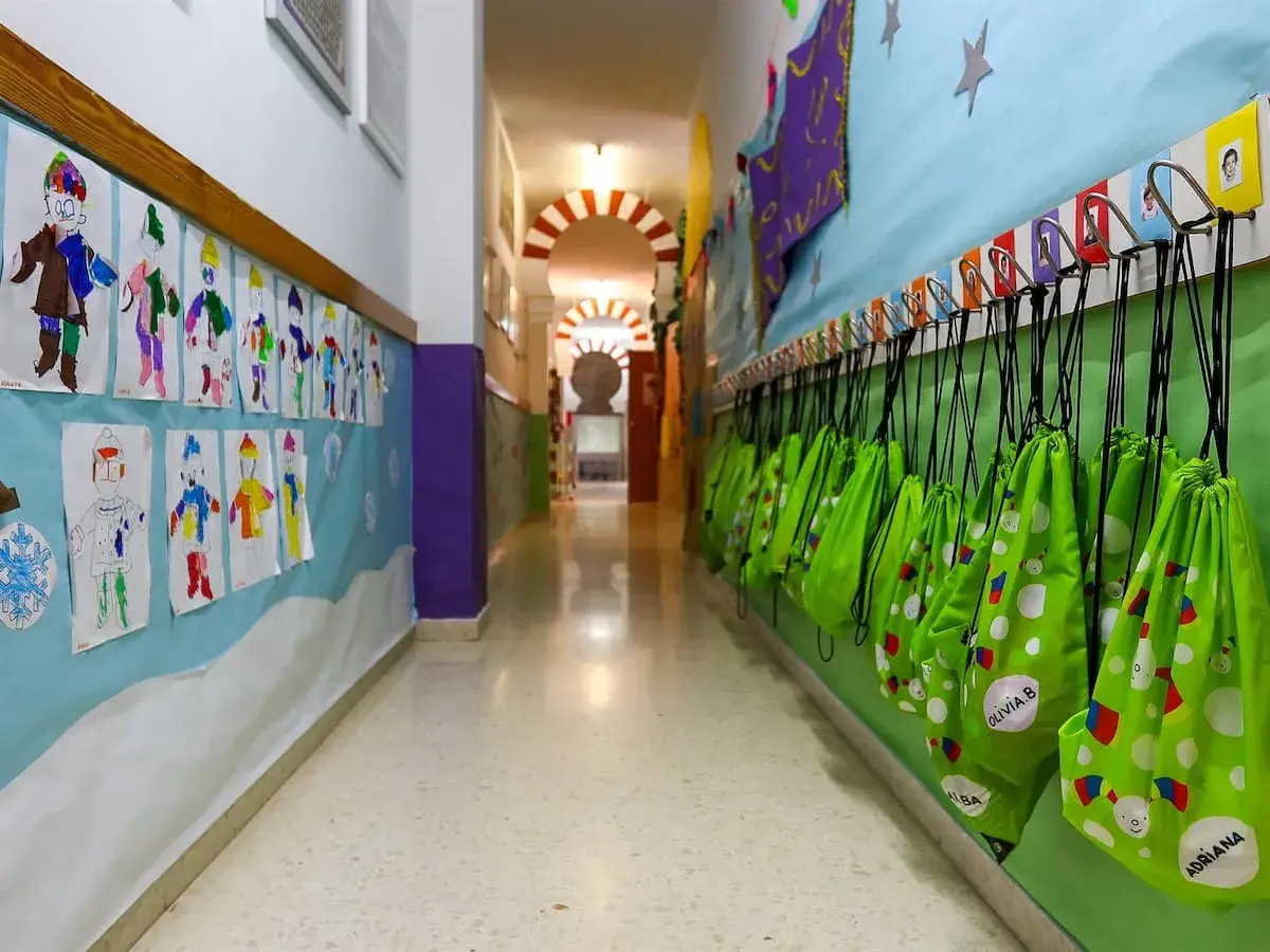 Pasillo de guardería en Comunidad Valenciana. Dibujos infantiles decoran la pared, y mochilas verdes cuelgan listas para la ayuda.