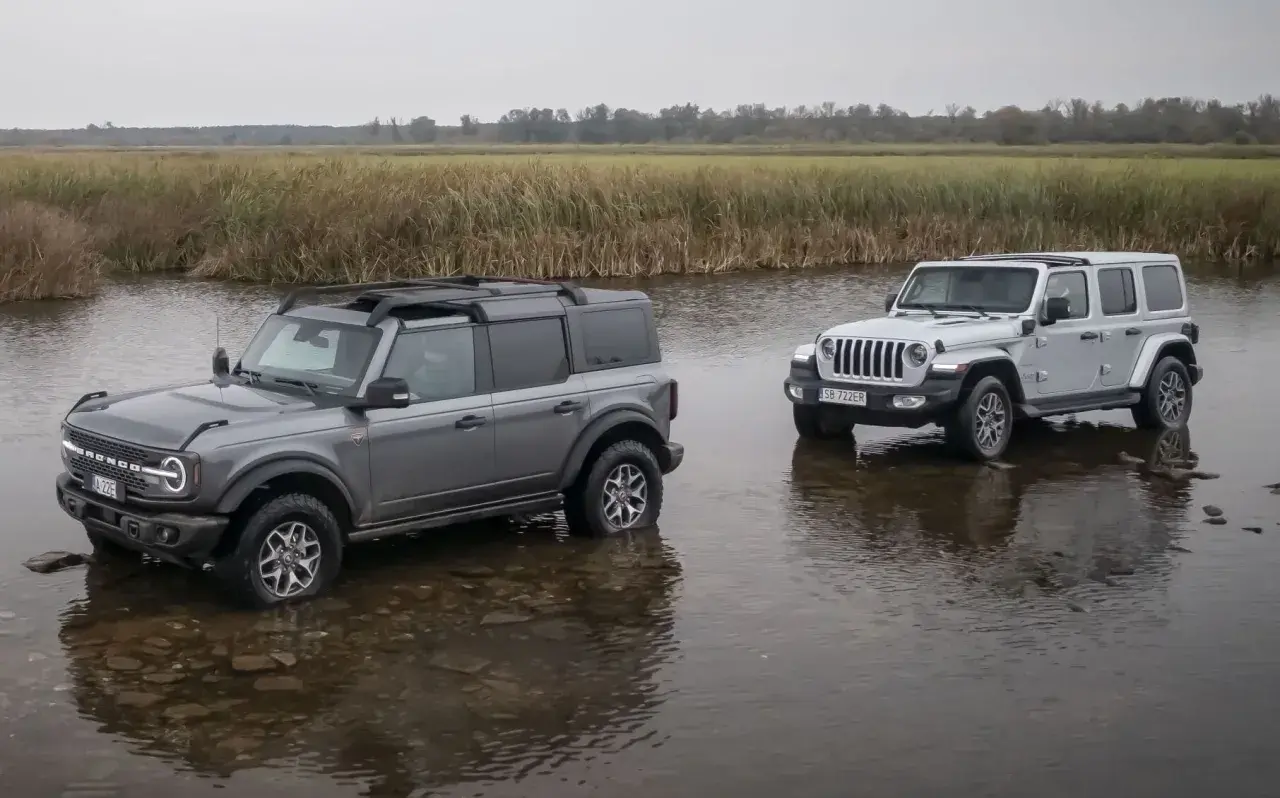 Szary Ford Bronco i biały Jeep Wrangler stoją w wodzie na tle łąki.