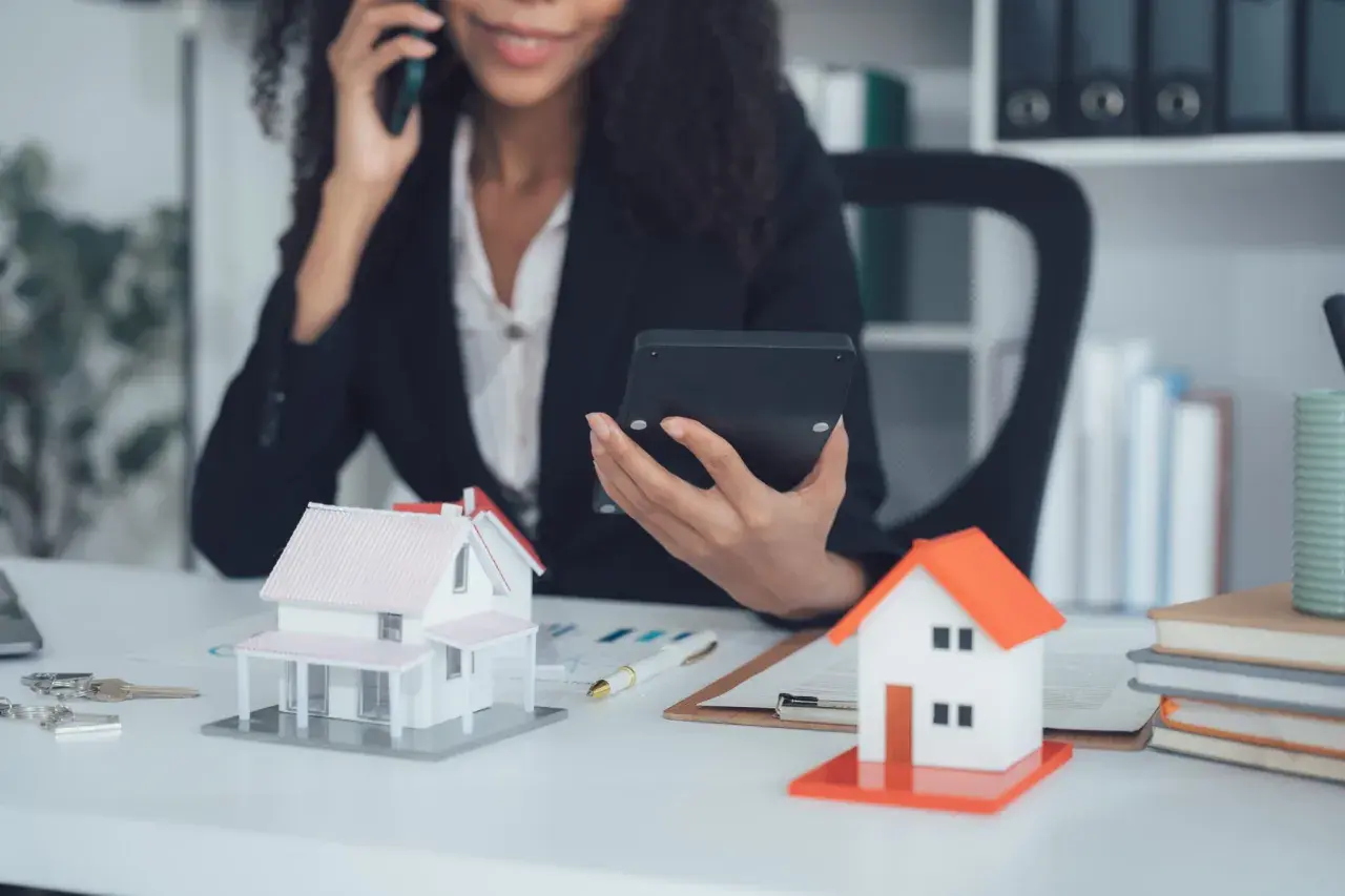 Agent immobilier au téléphone, calculatrice en main, devant des maquettes de maisons. Elle prépare une vente à terme.