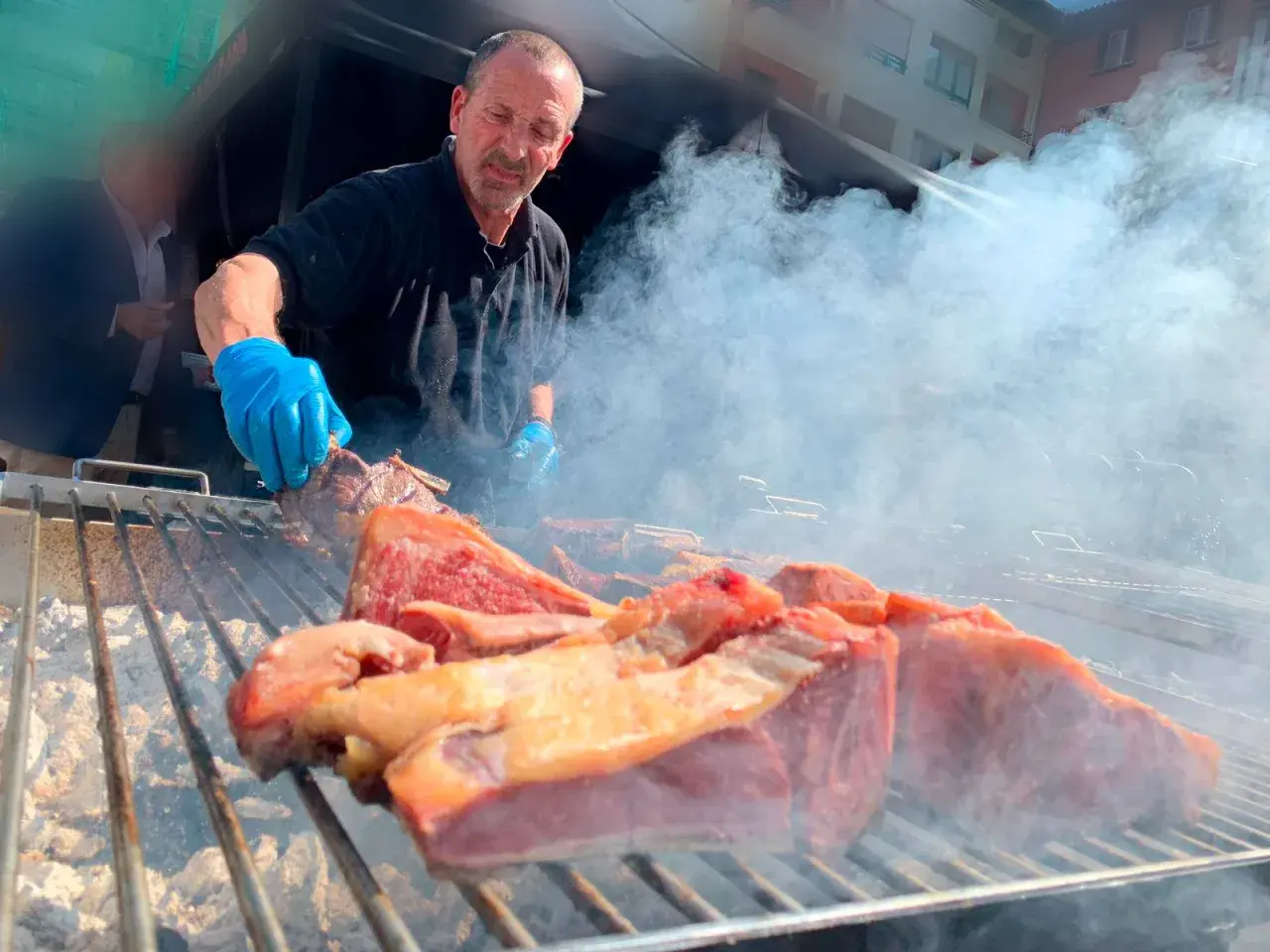 Hombre con guantes azules cocina carne en una parrilla humeante, un festín de carne en el **meat carnival Valencia**.
