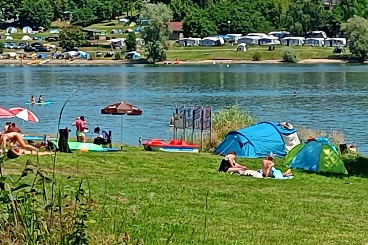 Letnie popołudnie nad jednym z malowniczych **jezior w Czechach**. Ludzie odpoczywają na trawie, opalają się, a w wodzie pływają kajaki. W tle widać pole namiotowe.