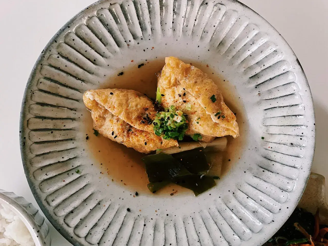 Two pieces of golden aburaage tofu simmer in a savory broth with leeks and seaweed, garnished with green onions and sesame seeds.