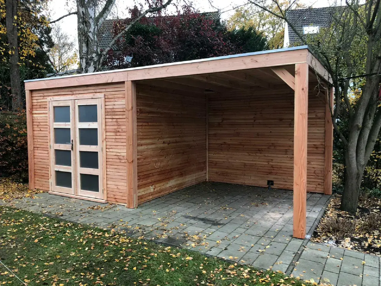 Neuer Holzschuppen mit Carport, das Dach ist mit Dachpappe verlegt. Herbstlaub liegt auf dem Boden.