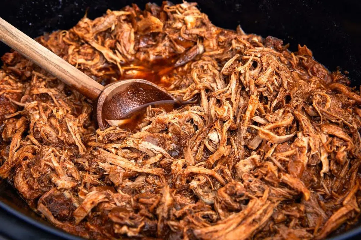En stor gryta med saftig, långkokt pulled pork i en mörk sås. En träsked vilar i den läckra pulled pork grytan.