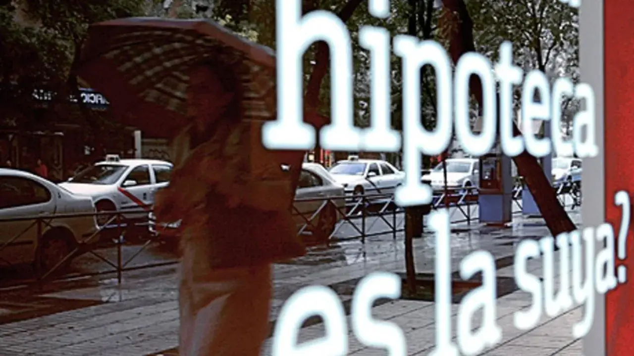 Reflejo en un escaparate: "Hipoteca, ¿es la fein suya?". Una persona con paraguas pasa por la calle lluviosa con taxis.