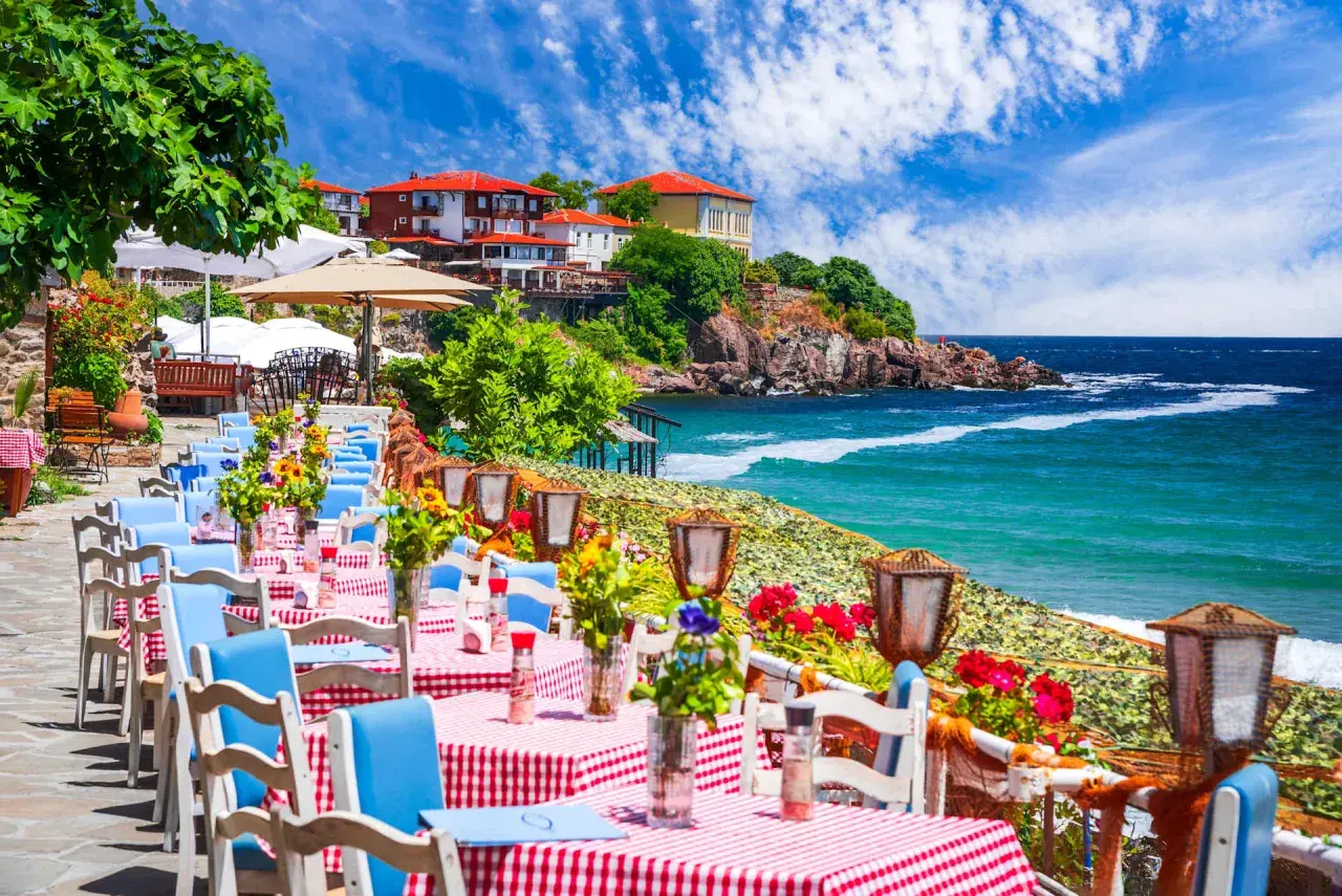 Urokliwa restauracja nad morzem w Bułgarii. Stoliki z czerwono-białymi obrusami i niebieskimi krzesłami czekają na gości, a w tle widać turkusowe morze i malownicze klify.