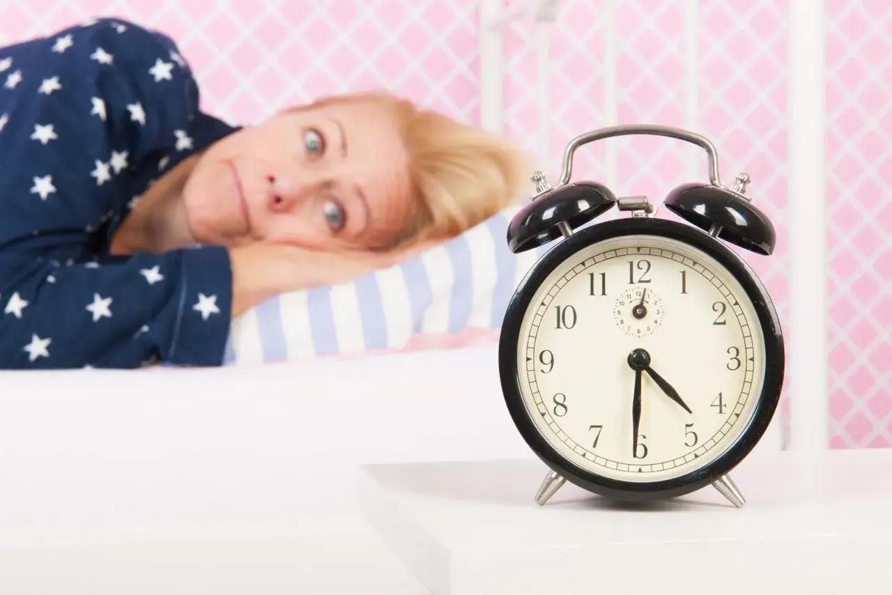 Mujer en pijama mira un despertador, su expresión refleja la **intención paradójica** de querer dormir pero no poder.