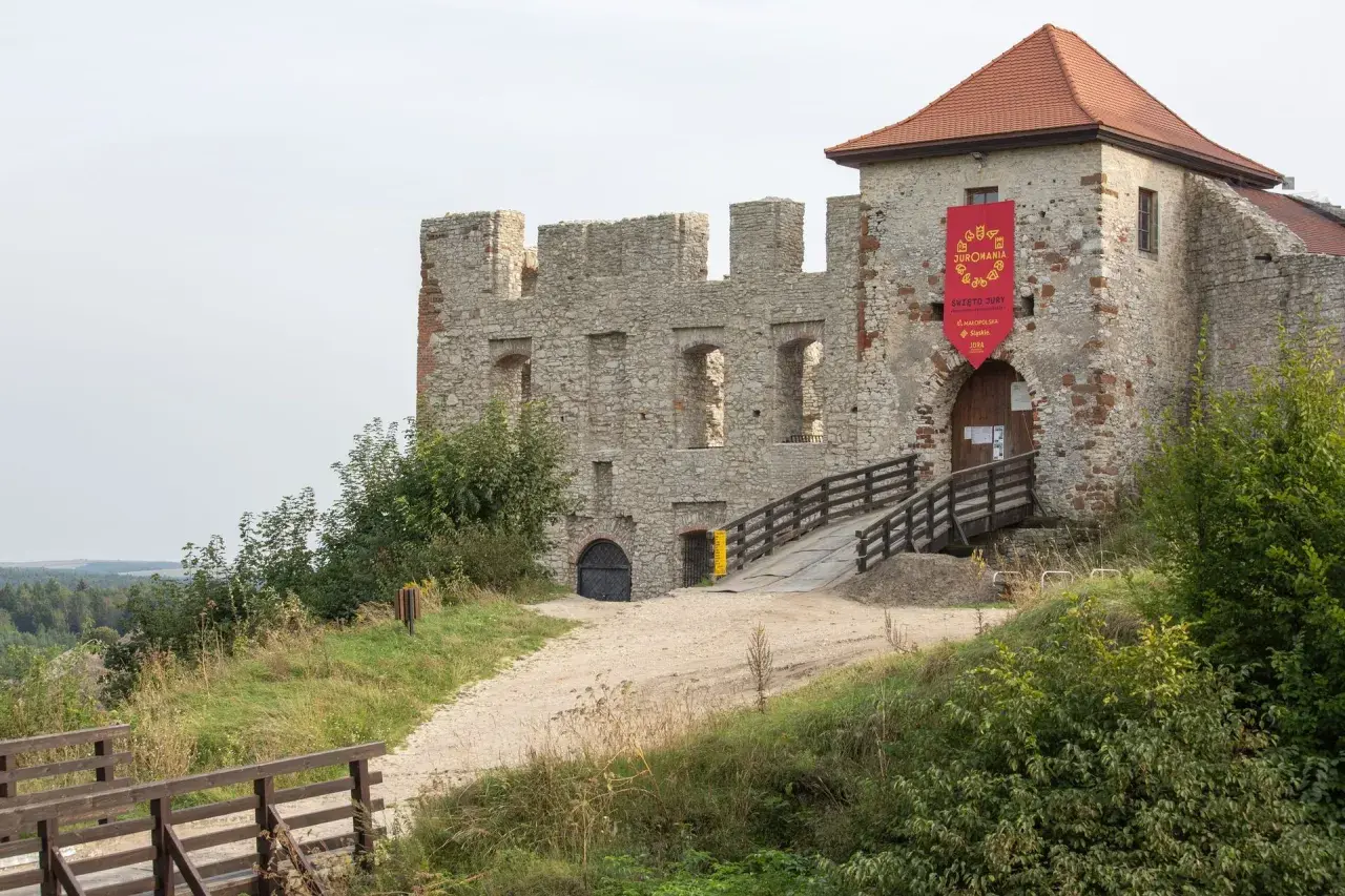 Kamienne mury zamku w Rabsztynie, z drewnianym mostkiem prowadzącym do bramy. Czerwony baner zaprasza na wydarzenie.