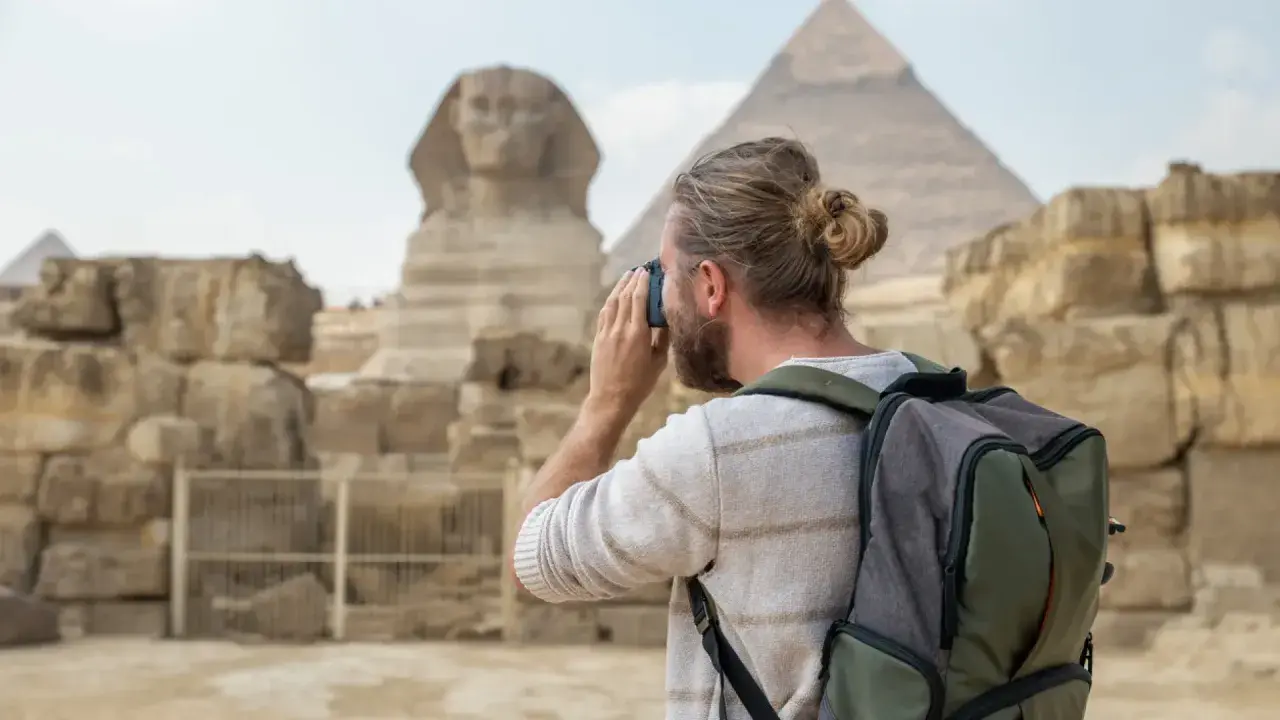 Hombre con mochila y cámara fotográfica admira la Esfinge y las pirámides. Es seguro viajar a Egipto y vivir esta experiencia.