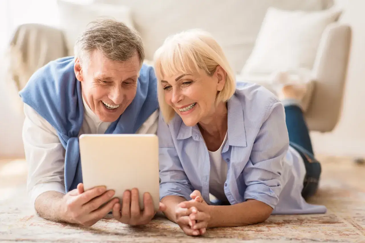 Ein lachendes älteres Paar liegt auf dem Boden und schaut auf ein Tablet. Ihre positiven senioren dates erfahrungen werden durch ihr Lächeln deutlich.