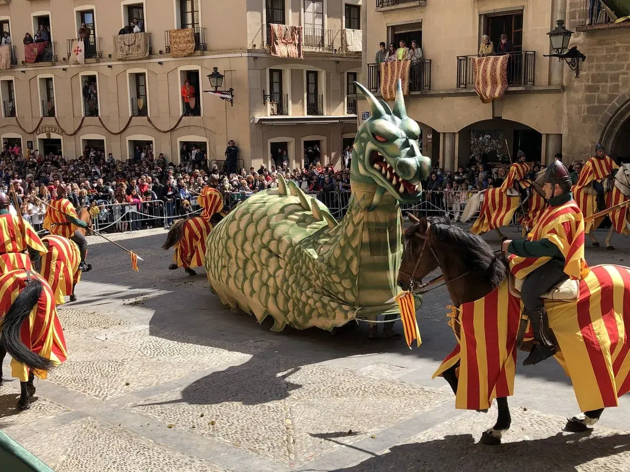 Un dragón gigante desfila en las fiestas de Alcañiz, acompañado por jinetes con trajes de rayas rojas y amarillas.