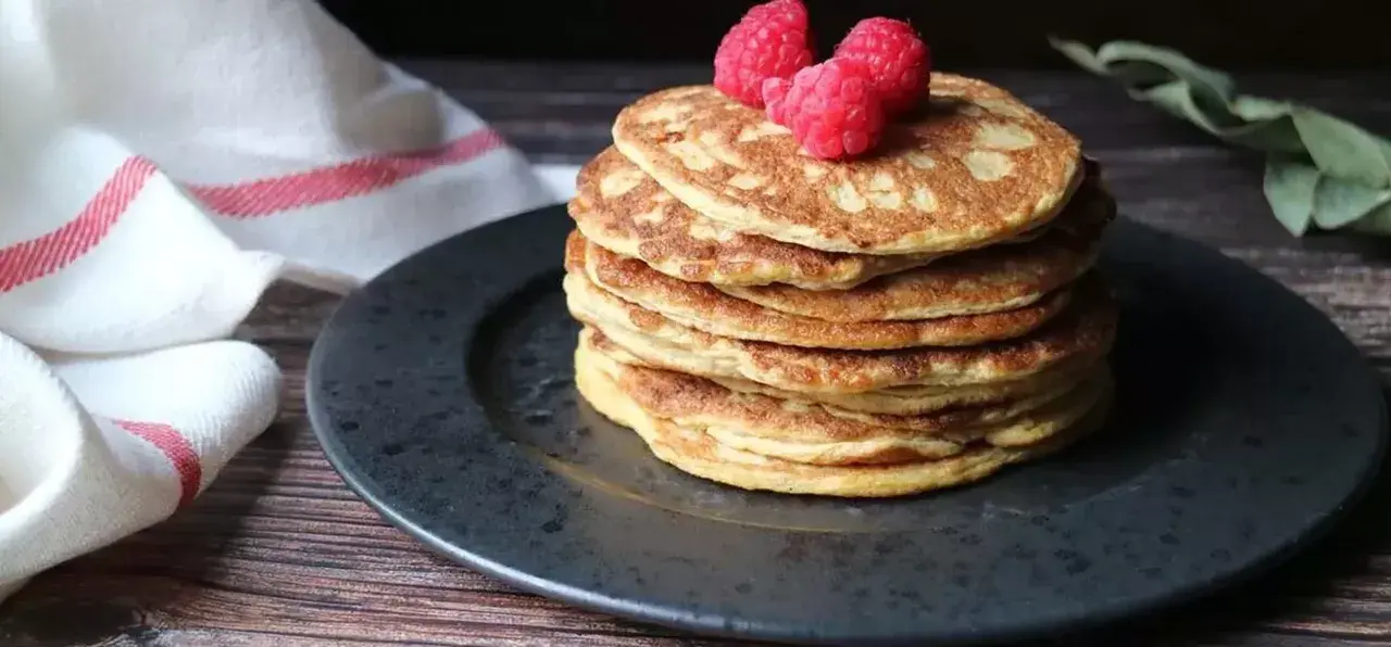 En hög med nyttiga pannkakor toppas med hallon, serverade på en mörk tallrik med en vit handduk med röda ränder i bakgrunden.
