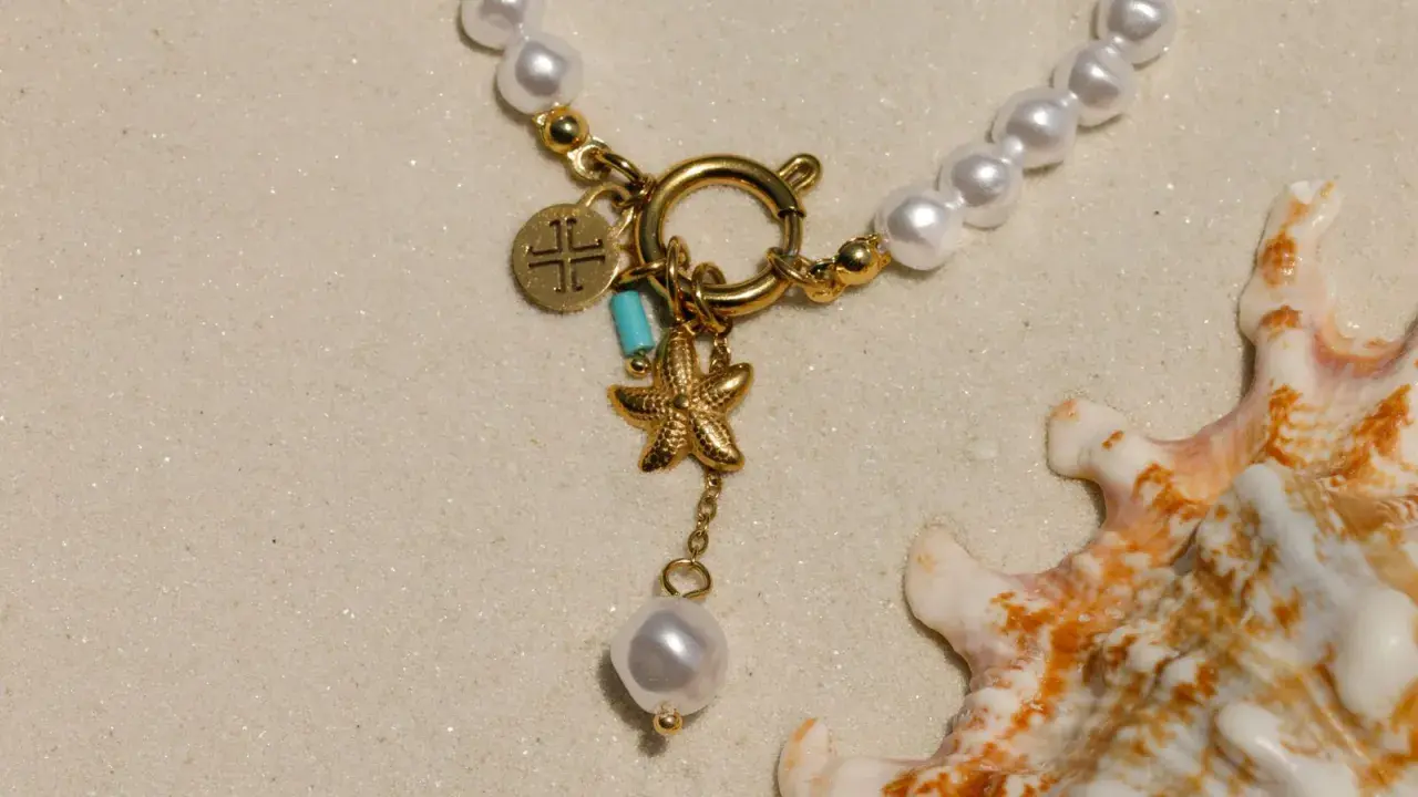 Un delicado llavero de oro con charms de estrella de mar y perla, sobre arena y junto a una concha marina.