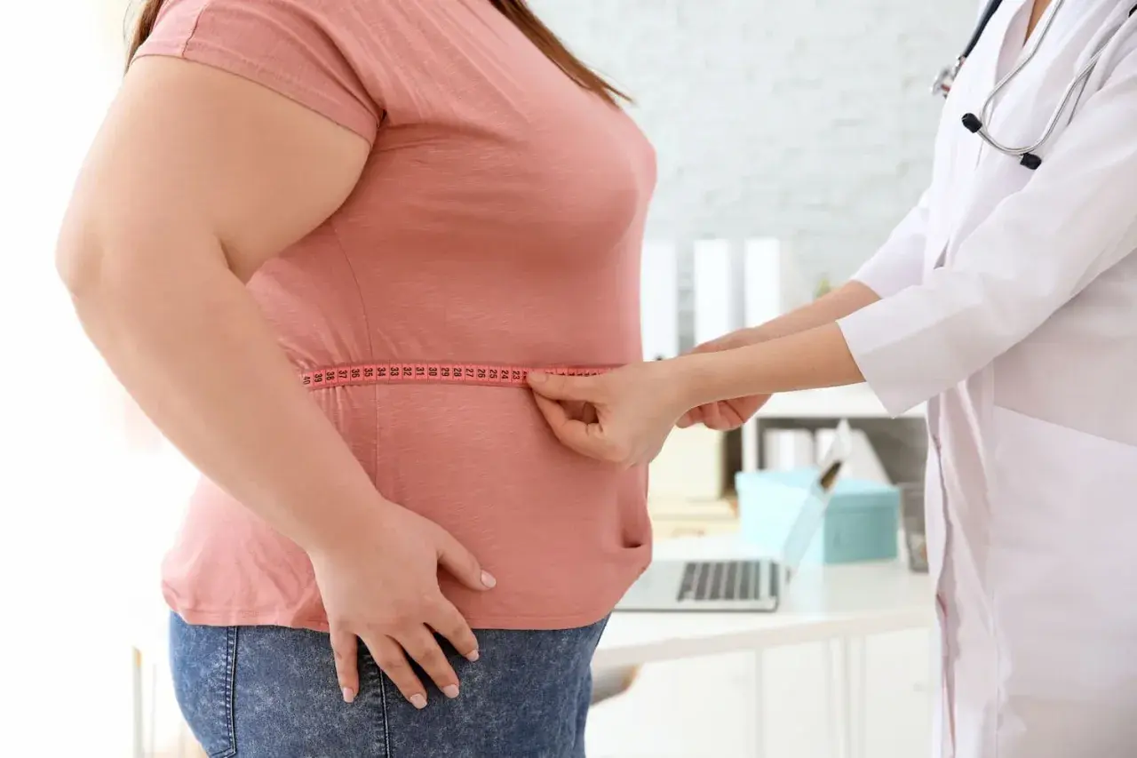 Arzt misst Taille einer übergewichtigen Frau. Mögliche Ursachen für Gewichtszunahme wie Eisenmangel werden besprochen.