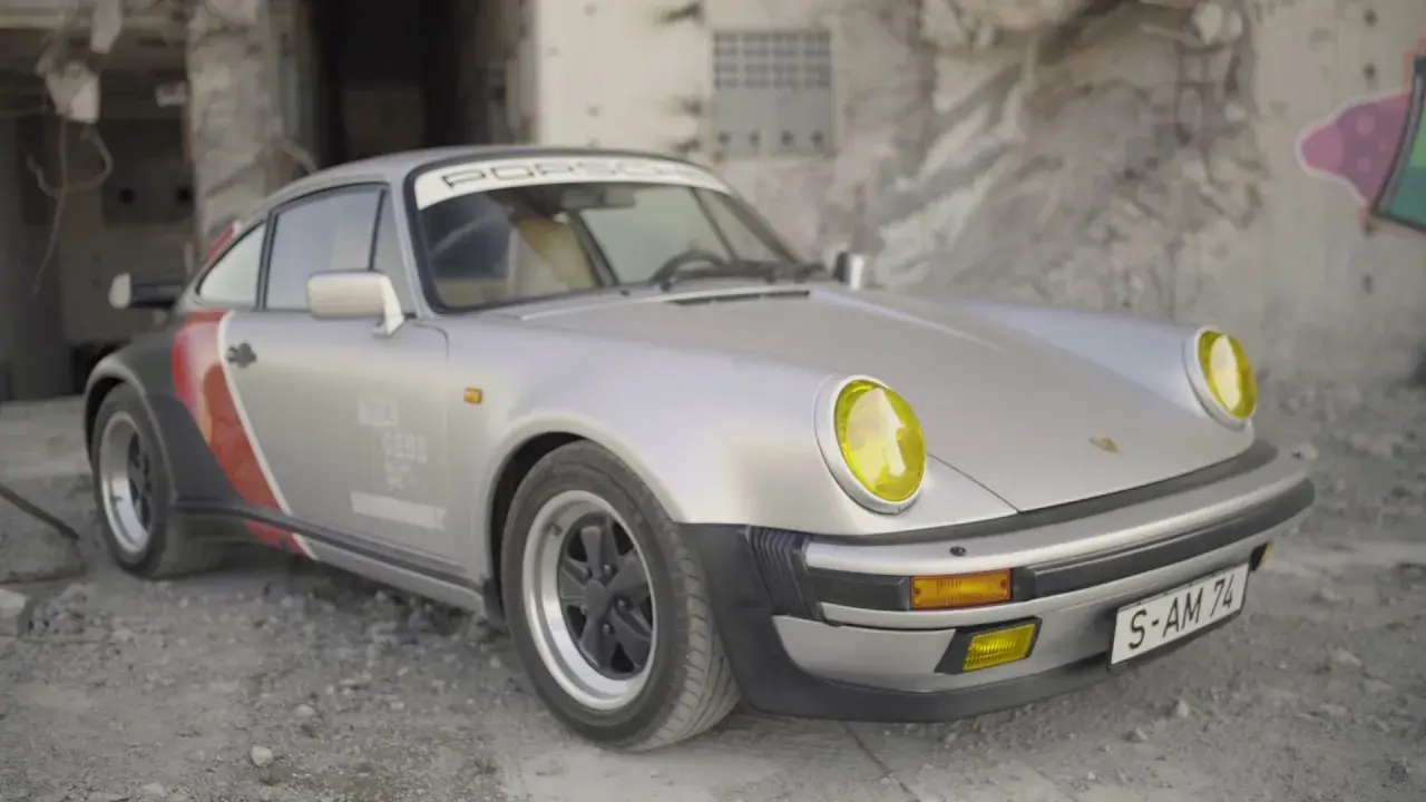 Srebrny Porsche 911 z żółtymi reflektorami, w stylu cyberpunk, zaparkowany na tle zniszczonej ściany z graffiti.