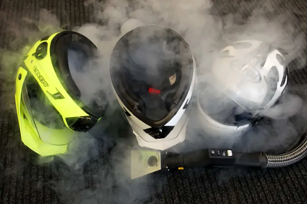Tres cascos de moto rodeados de vapor. Uno amarillo brillante, uno blanco y negro, y uno blanco con diseño. Un limpiador a vapor está cerca. ¿Son estas las peores marcas de cascos para moto?