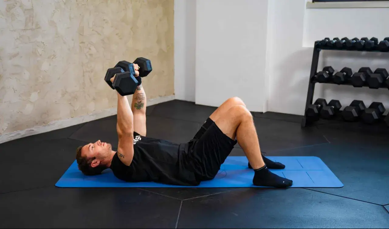Uomo esegue esercizi pettorali con manubri su tappetino blu. Allenamento in palestra con rack di pesi sullo sfondo.