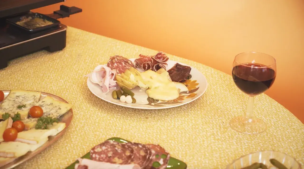 Un verre de vin rouge accompagne une raclette savoureuse, des charcuteries et des fromages sur une table jaune.