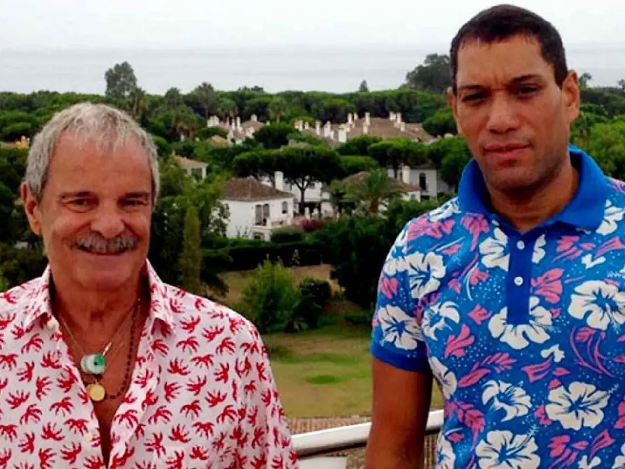 Elio Valderrama Prescott y un amigo posan con camisas coloridas frente a un paisaje de casas y árboles.