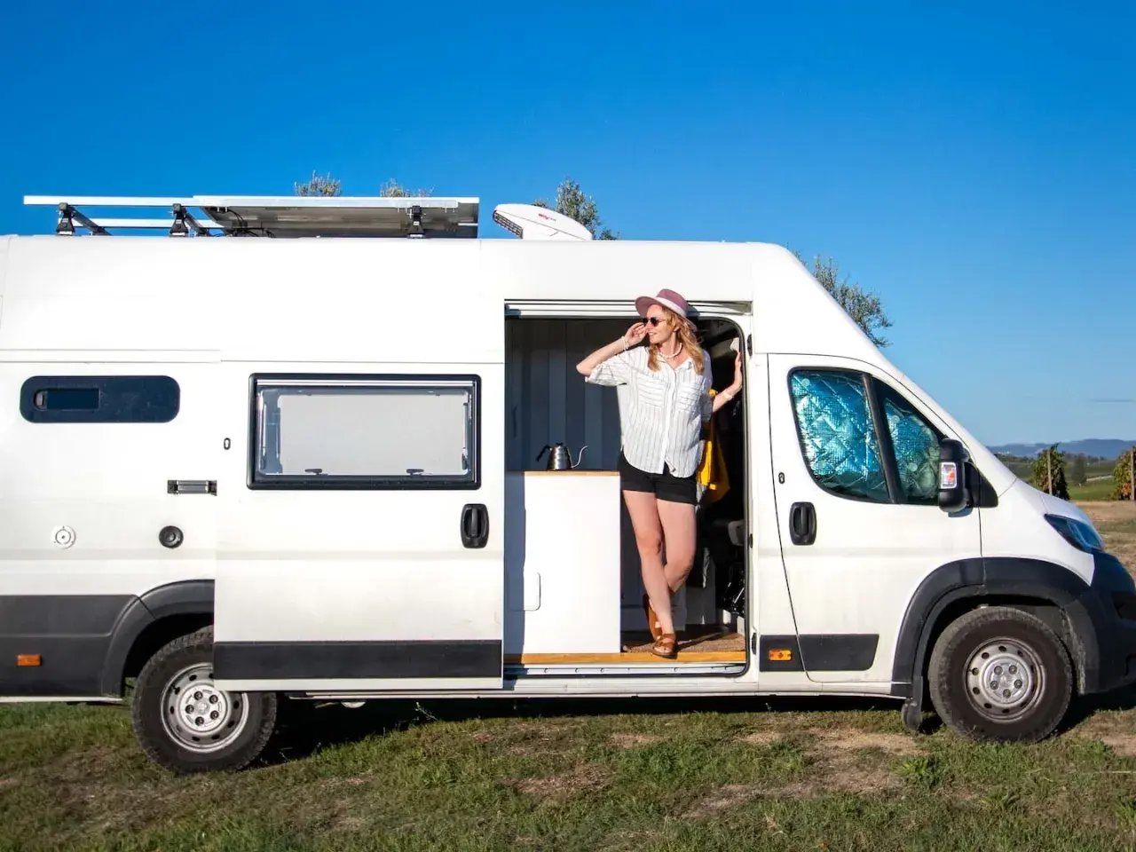 Kobieta w kapeluszu wychodzi z białego kampera z panelem słonecznym na dachu. Jej camper system jest gotowy na przygodę.