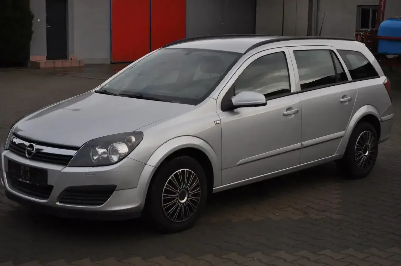 Srebrny Opel Astra H 1.6 benzyna, kombi, zaparkowany na kostce brukowej przed budynkiem z czerwonymi drzwiami.