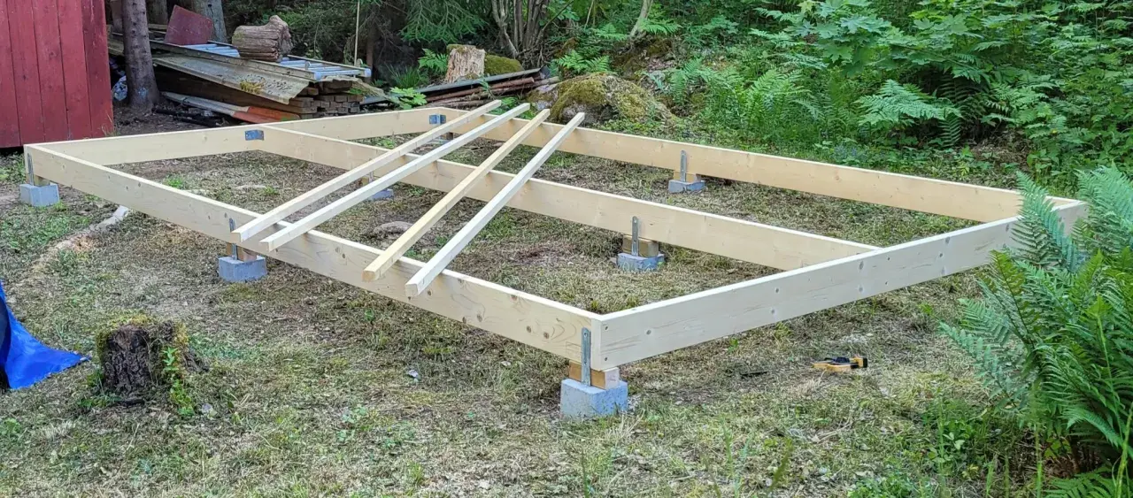 Grundläggande stomme för att bygga hus på plintar. Träbjälkar vilar på betongblock i en skogsmiljö.
