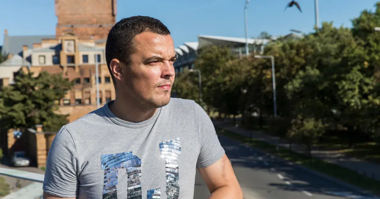 Dawid Janczyk w szarej koszulce z błyszczącym napisem, na tle miejskiego krajobrazu z budynkami i drzewami.