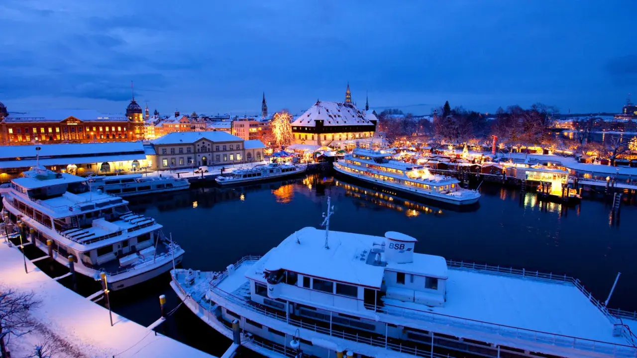 Schneebedeckte Schiffe im Hafen von Konstanz, beleuchtet vom Glanz des Weihnachtsmarktes.