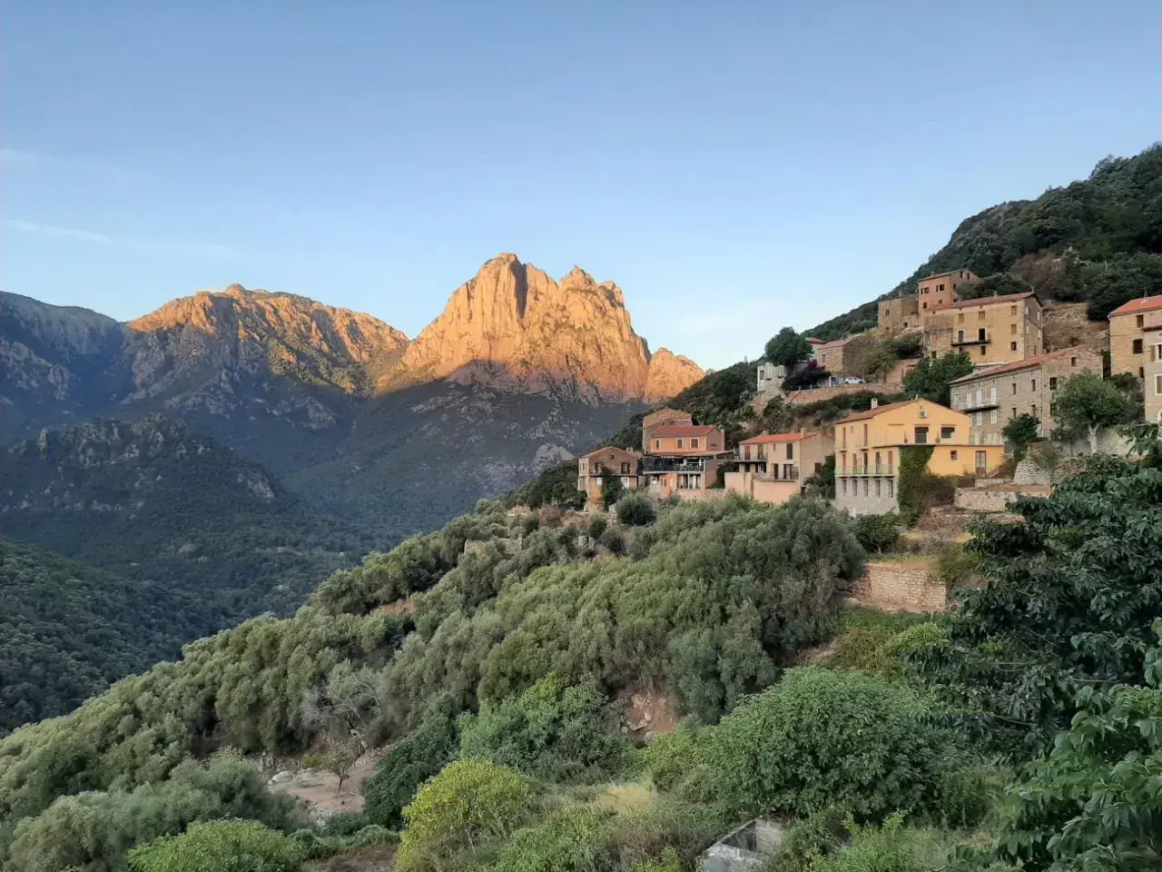 Village perché sur une colline, baigné par le soleil couchant. Les montagnes du nord, d'une difficulté certaine, se dressent en arrière-plan.