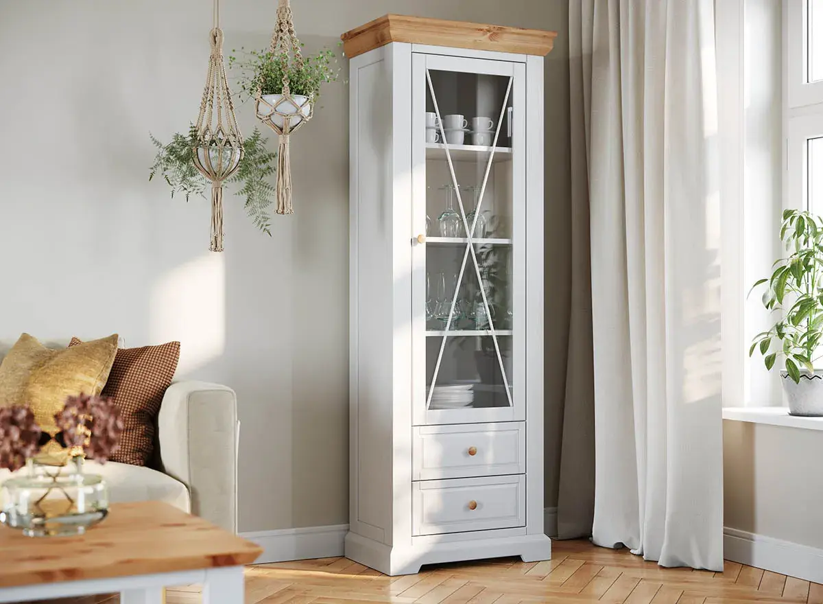Jasny salon biało drewniany z witryną pełną szkła i porcelany. Wiszące makramy z roślinami dodają przytulności.