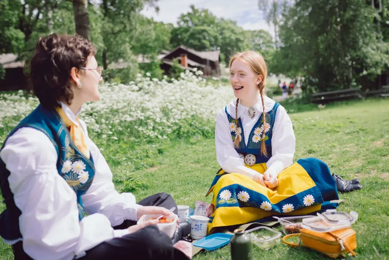 Två personer i folkdräkter njuter av en picknick under midsommar på Skansen.