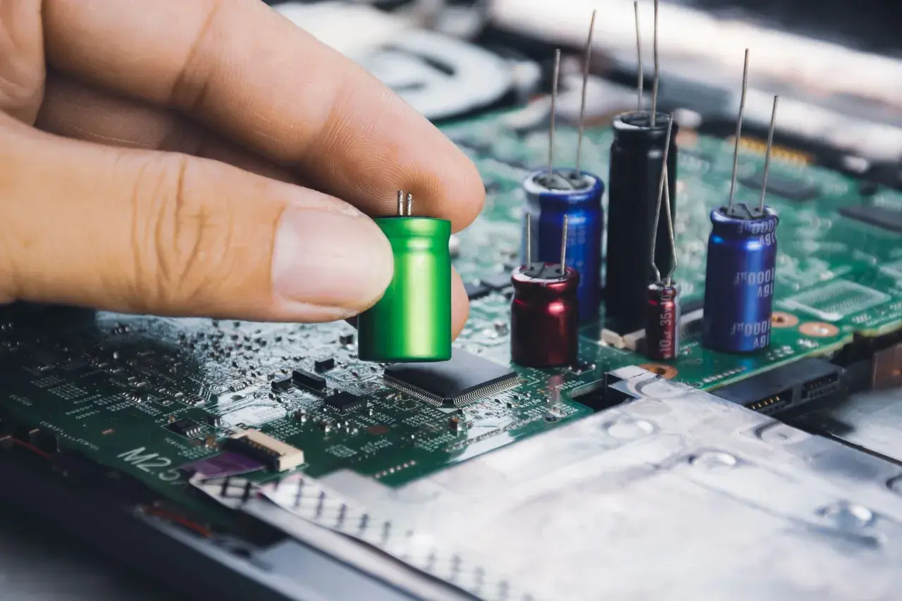 Dłoń trzyma zielony kondensator, przygotowując go do montażu na płycie głównej. W tle widać inne kondensatory i układy elektroniczne.