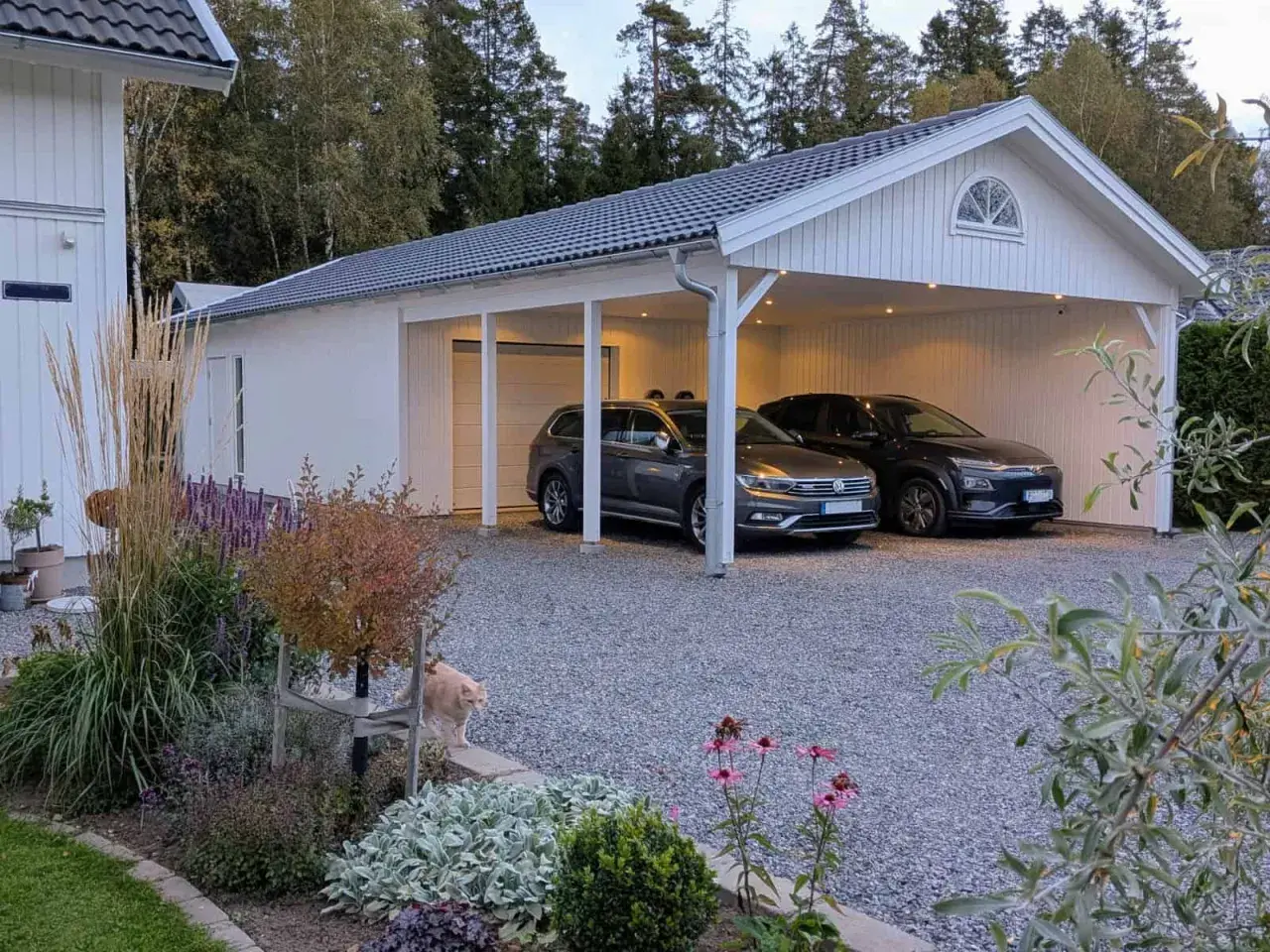 Vitt garage med två bilar under taket. En katt smyger i rabatten framför. Perfekt för att bygga garageport.