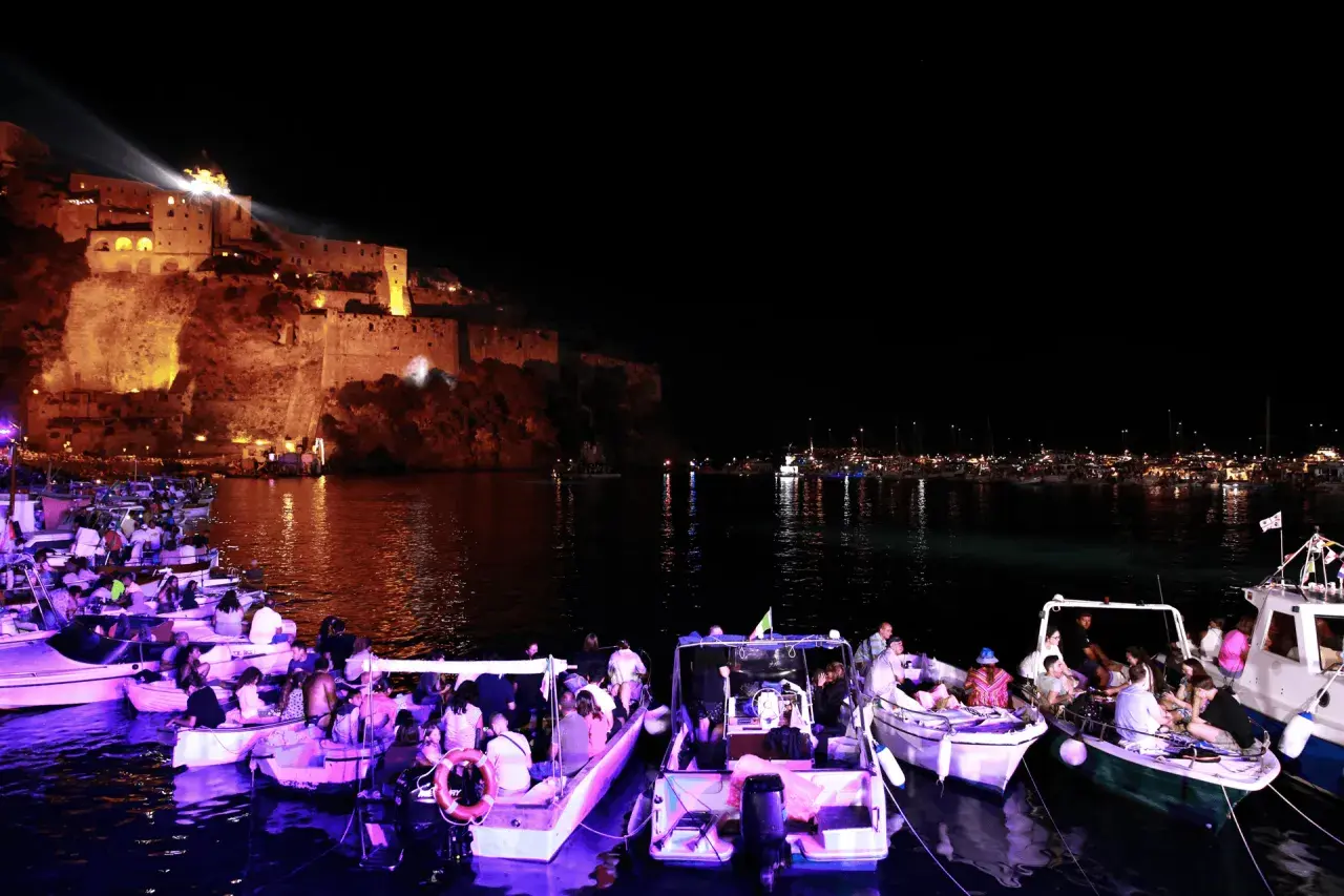 Una magica festa a Ischia: barche illuminate e gente festante con il castello illuminato sullo sfondo.