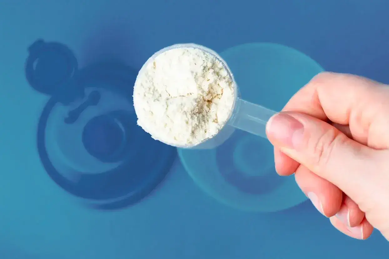 Una mano sostiene una cuchara con polvo blanco, ¿cuanta creatina tomar? Fondo azul con silueta de batidora.