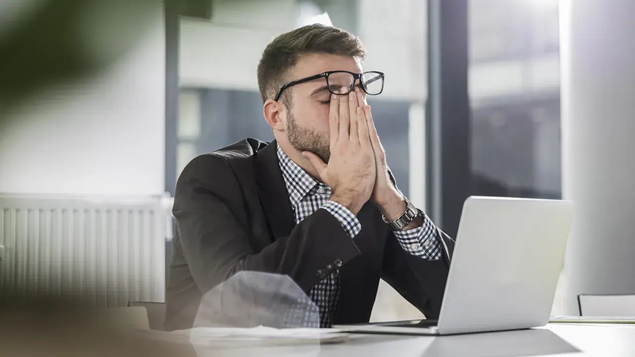 Man med glasögon sitter vid en laptop, gnuggar ögonen. Ett klassiskt dilemma exempel på stressad arbetsdag.