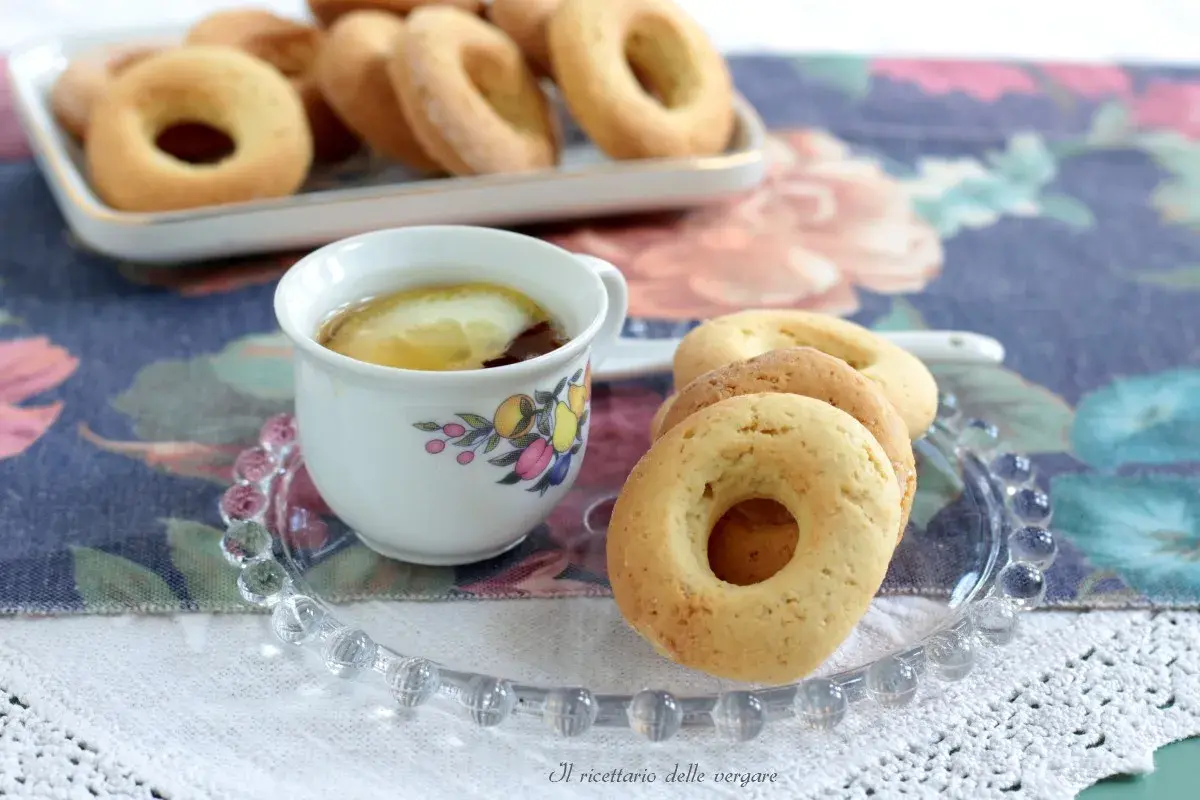 Biscotti senza burro dorati su un piattino di vetro, accanto a una tazza di tè con limone.