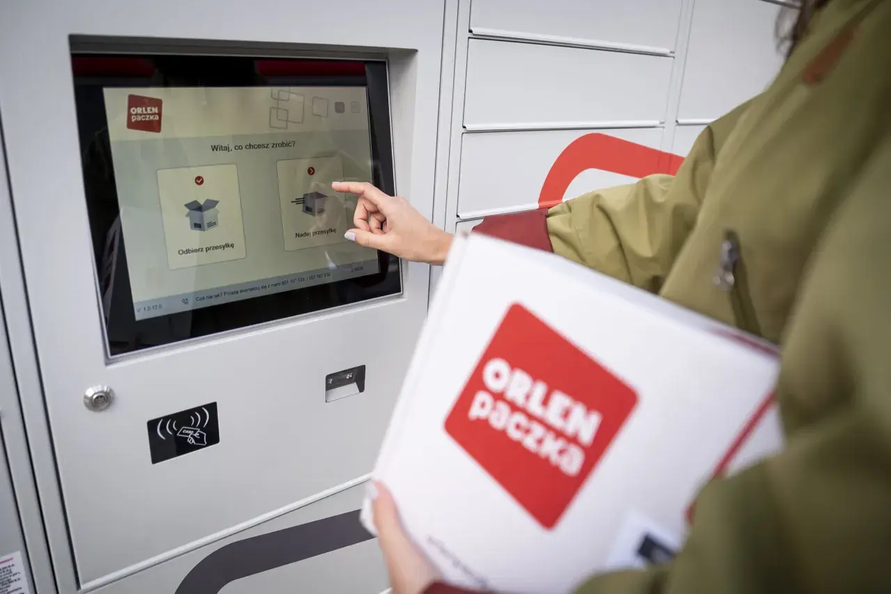 Kobieta z paczką ORLEN PACZKA dotyka ekranu automatu, wybierając opcję nadania przesyłki.