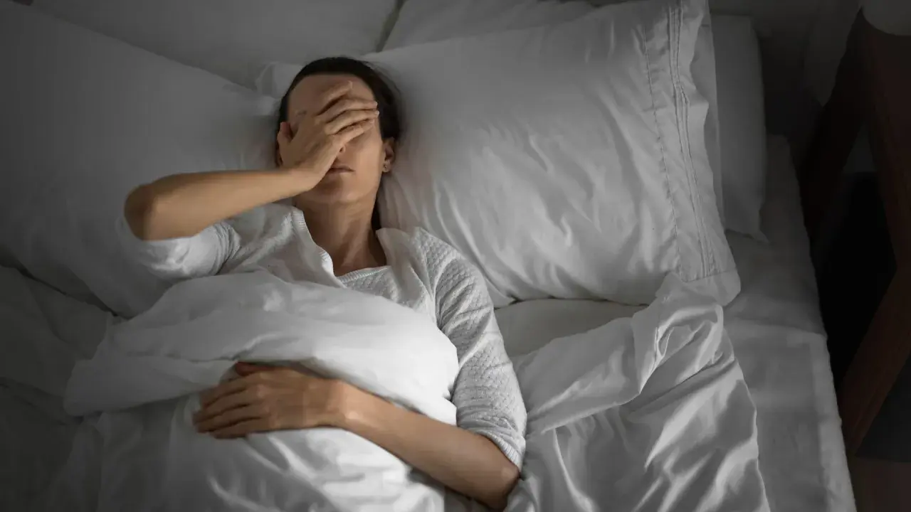 Frau liegt im Bett, bedeckt mit einer Decke, und hält sich die Hand vor die Augen. Sie kann nicht schlafen.