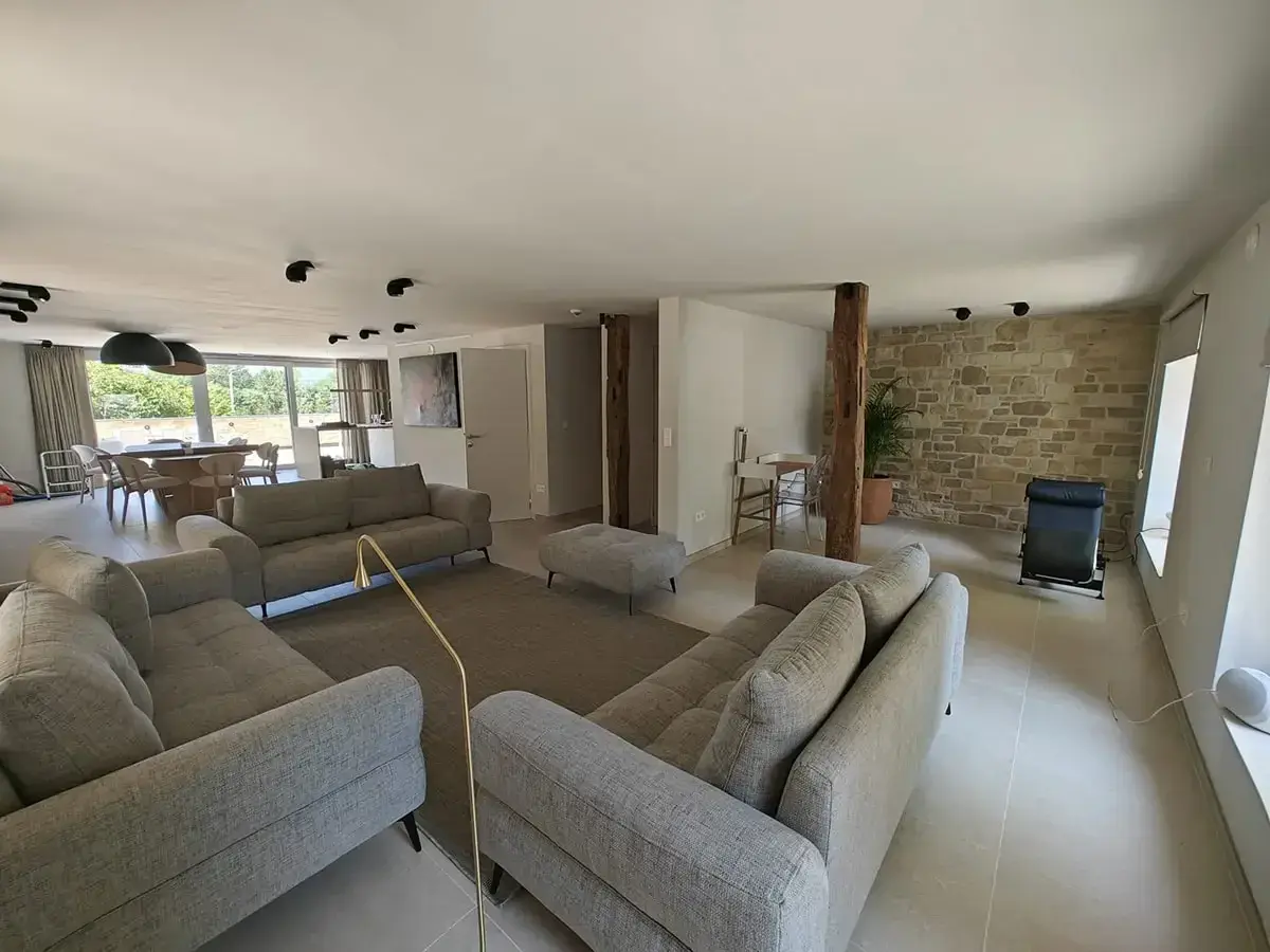 Salon moderne avec canapés gris, un pouf, un tapis, et un mur en pierre apparente rustique. Une plante verte et un fauteuil noir complètent l'espace.