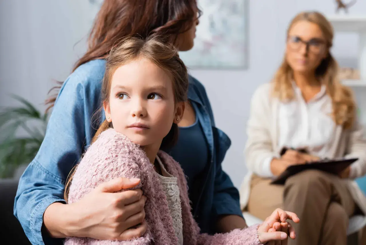 Dziewczynka w ramionach matki, zaniepokojona, szuka wsparcia. W tle terapeuta. Obrazuje skutki niepraktykowania wiary przez rodziców dla duchowości dziecka.