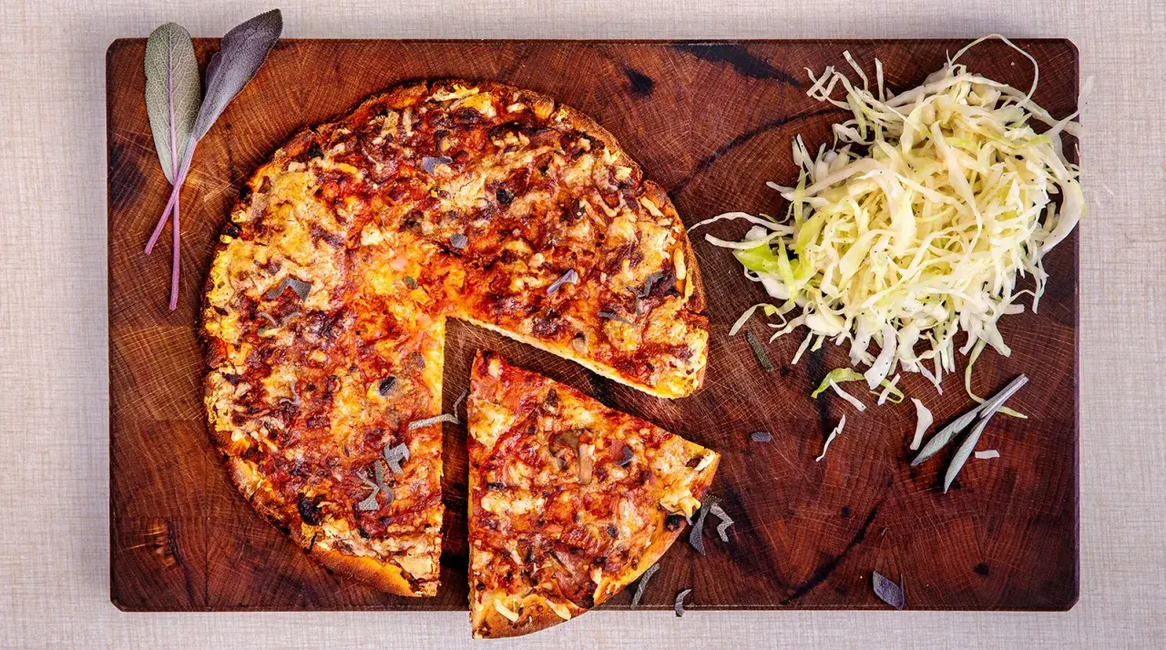 En hel och en skivad pizza på en träskärbräda, med en hög strimlad kål. En klassisk kombination som ger en hint om pizzasallad ursprung.