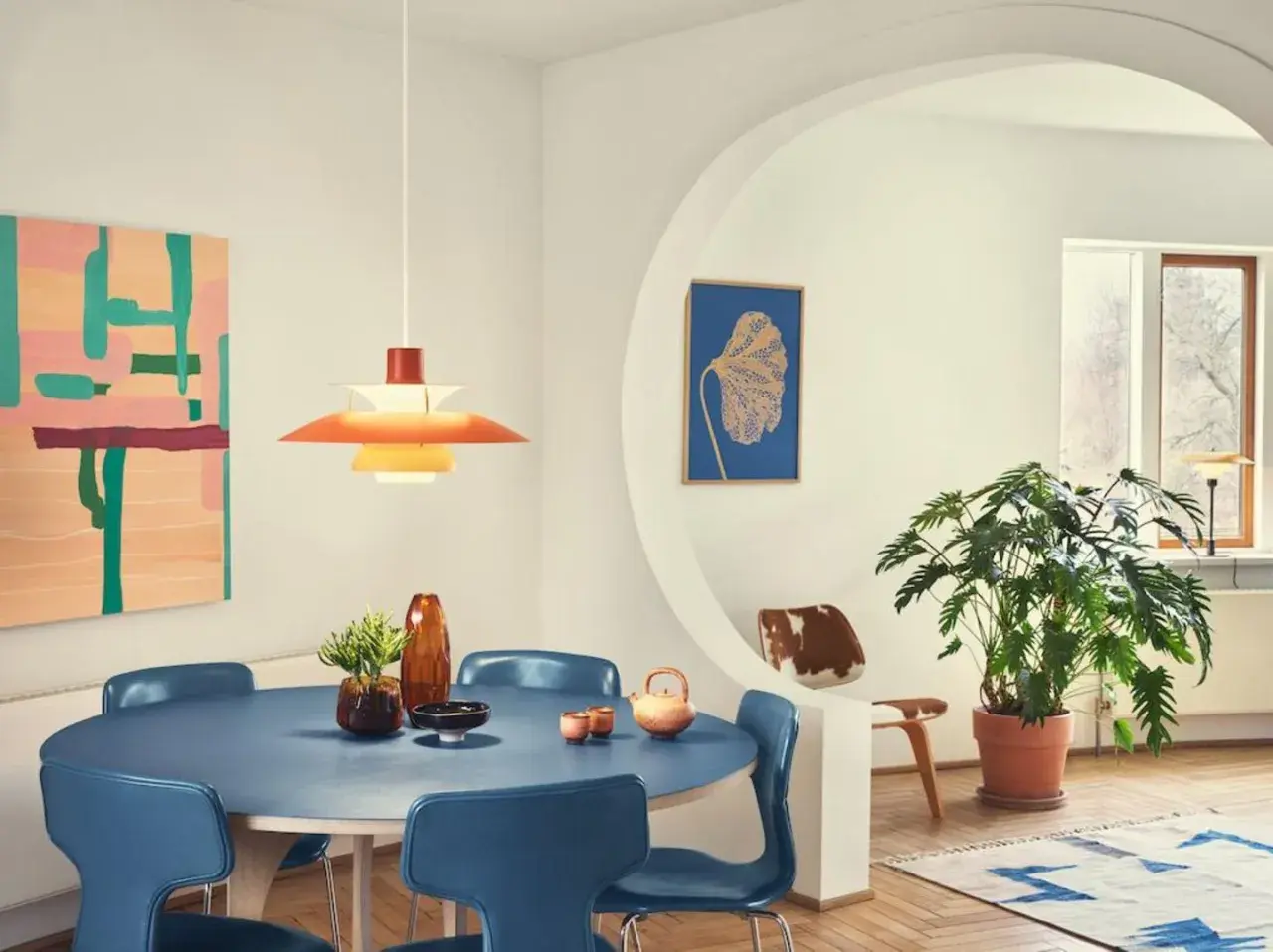 Ein Esszimmer im **midcentury modern** Stil mit blauem Tisch, blauen Stühlen, einer bunten Wandkunst und einer markanten Hängeleuchte.