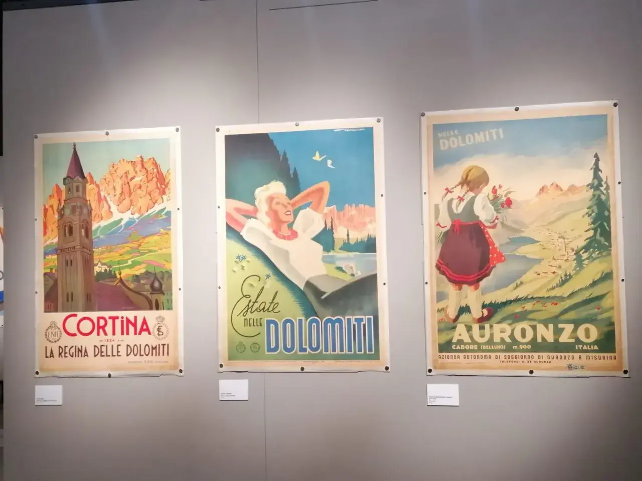 Tre manifesti vintage di una collezione salce promuovono le Dolomiti: Cortina, una donna rilassata e una bambina con fiori ad Auronzo.