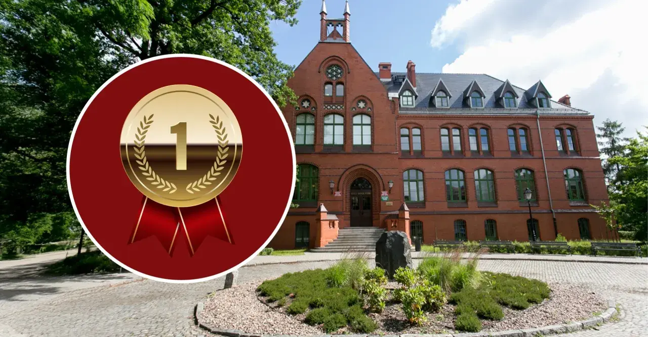 Złoty medal z cyfrą "1" na tle ceglanego budynku i zieleni.