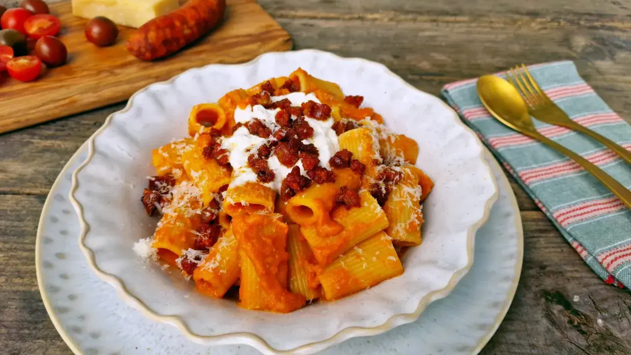Un plato de macarrones con chorizo, cubiertos con queso rallado y una cucharada de crema. Al fondo, tomates y un trozo de queso.