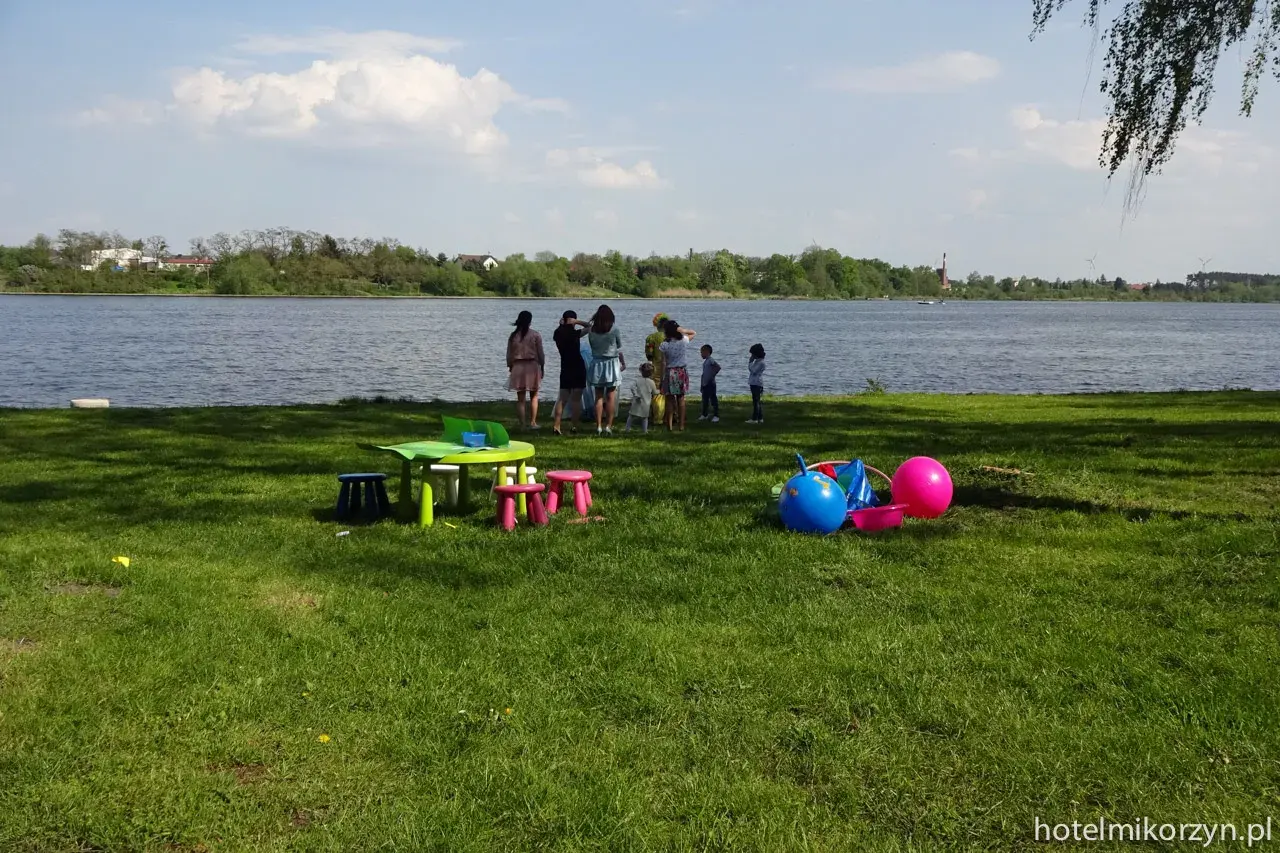 Dzieci bawią się nad jeziorem Mikorzyńskim. Na trawie kolorowe zabawki: piłki, stolik i krzesełka.
