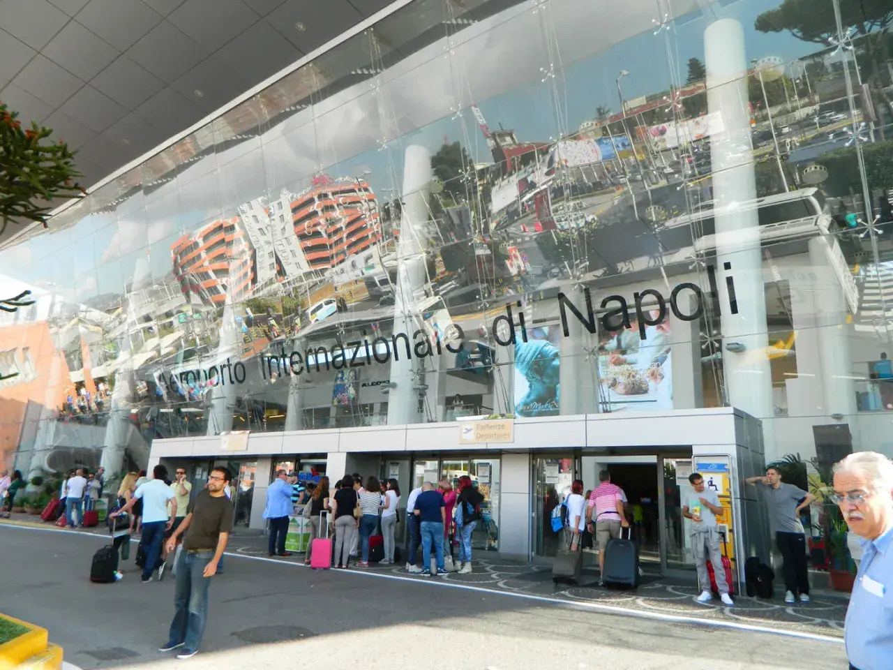 Ludzie z bagażami przed nowoczesnym budynkiem Aeroporto Internazionale di Napoli.