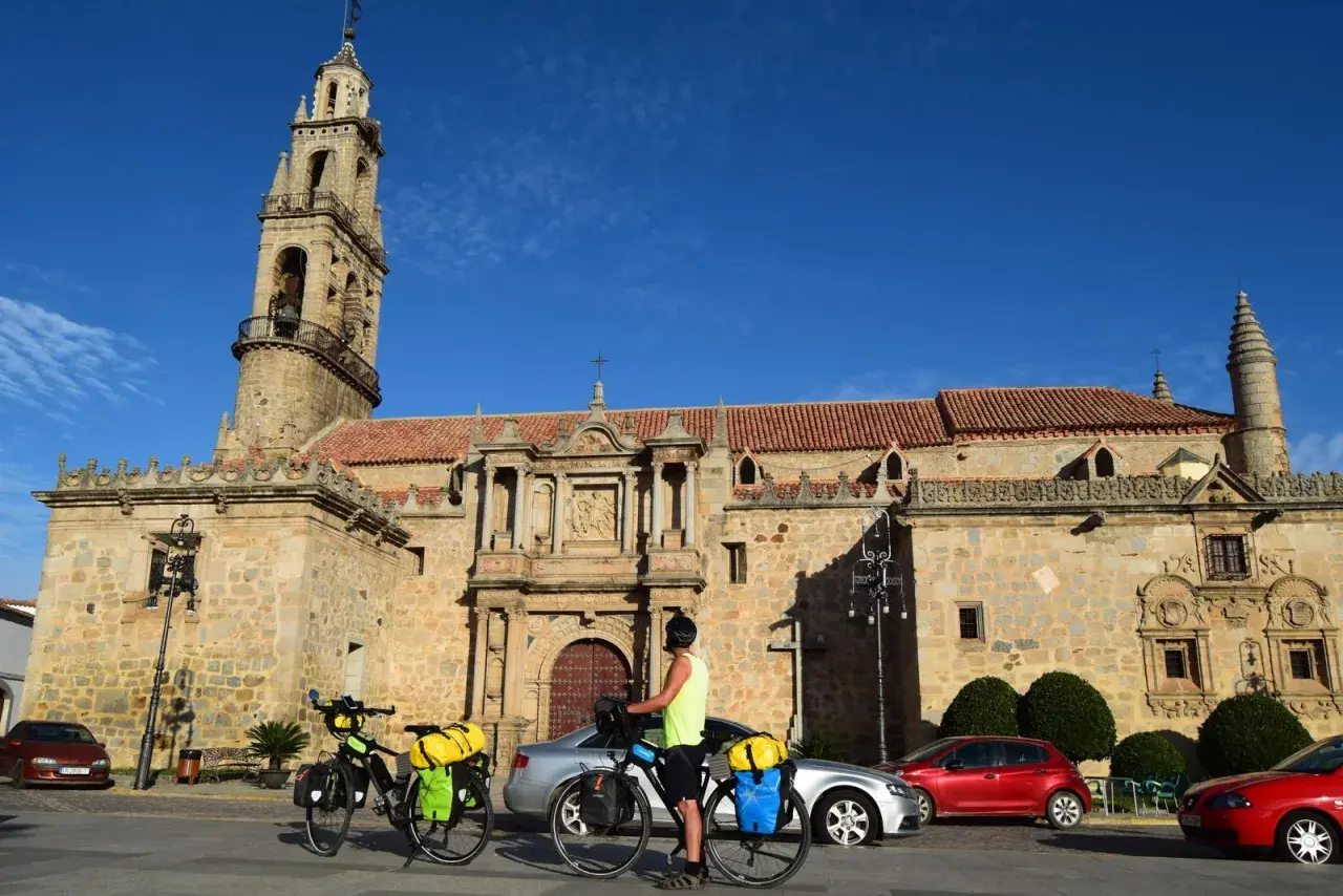 Ciclistas preparan sus bicicletas para continuar las etapas de Transandalus frente a una iglesia histórica bajo un cielo azul.